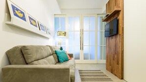 A living room with a sofa, a TV mounted on a wooden panel, and a sliding glass door leading to a balcony. - Side sea View 3min From the Beach Pj204 (Rio de Janeiro)