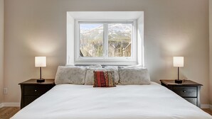 A bedroom with a large bed, two bedside tables with lamps, a window with a mountain view, and a patterned pillow. - RATES REDUCED! Dogs Welcome! Stunning Views & Unbeatable Location! MainC (Breckenridge)
