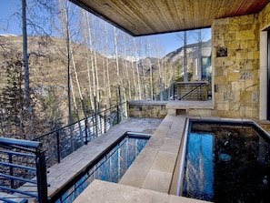 Ein moderner Außenpoolbereich mit Steinwänden und einer Holzbalkendecke, umgeben von einer Steinterrasse und Metallgeländern, mit Blick auf eine bewaldete Berglandschaft.