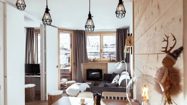 Ein gemütliches Wohnzimmer mit Kamin, Holzmöbeln und Blick auf eine verschneite Landschaft durch große Fenster.