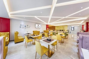 A modern dining area with yellow and purple seating, a visible restaurant section, and a ceiling with geometric lighting.