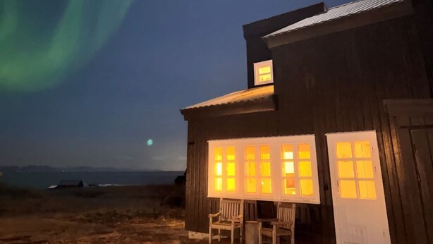 A wooden building with a porch and two chairs, under a night sky with the aurora borealis. - Panoramic Ocean & Glacier Views | Northern Lights Cottage | Hellnar, Iceland (Hellnar)