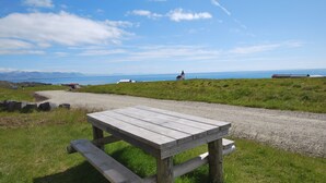 A wooden picnic table on a gravel path with grassy areas and a view of the sea and distant mountains. - Panoramic Ocean & Glacier Views | Northern Lights Cottage | Hellnar, Iceland (Hellnar)