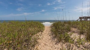 Un sentier de sable qui longe la plage et mène à l’océan, avec une végétation clairsemée de part et d’autre.