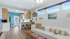 Un salon moderne avec un canapé blanc, une table basse en bois et un coin cuisine doté de meubles en bois et d’un plan de travail blanc.