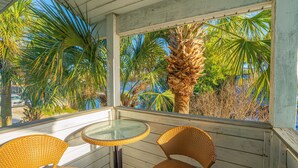 Un balcon abrité, doté de fauteuils en osier, d’une table en verre et offrant une vue sur des palmiers et un ciel bleu.