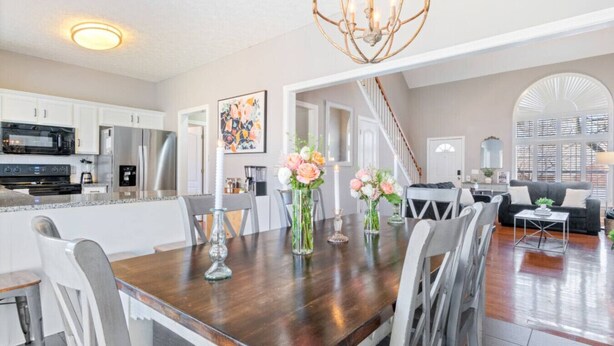 Een moderne keuken met witte kasten, een zwarte magnetron en een roestvrijstalen koelkast. Er is een eethoek met een houten tafel, witte stoelen en bloemige decoraties. Een kroonluchter hangt aan het plafond en een trap leidt naar een hoger niveau.