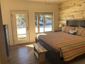 Quarto com uma cama grande, um banco, uma porta que dá para o exterior e uma janela com vista para árvores.