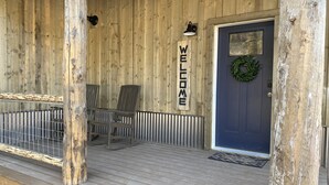 Une véranda en bois avec une pancarte « BIENVENUE », deux chaises et une porte ornée d’une guirlande.