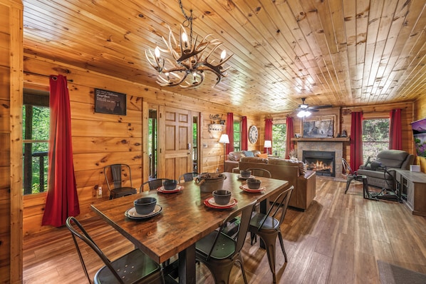 A wooden cabin interior with a dining table, chairs, a fireplace, and a ceiling fan.