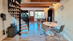A wooden staircase with a black railing leading to a second-floor area. A round table with chairs is positioned in the foreground, and a brick fireplace is visible in the background. - Lakefront Cottage Near Crystal Mountain & Rainier (Sumner)