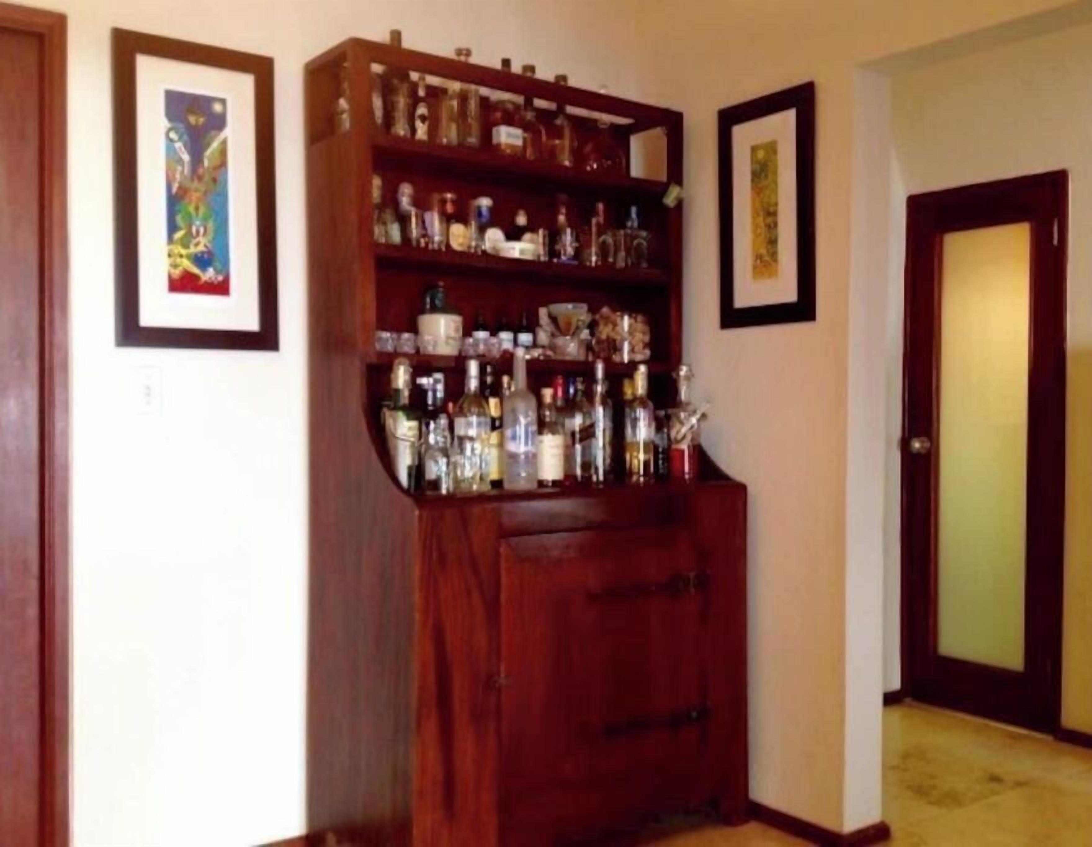 A wooden cabinet with shelves filled with various bottles, likely containing alcohol, in a room with two framed artworks on the walls.