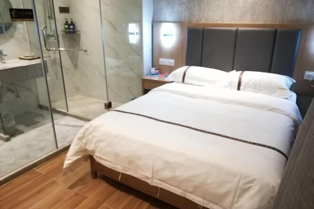 A hotel room with a bed, a glass shower enclosure, a bedside table with toiletries, and a headboard.