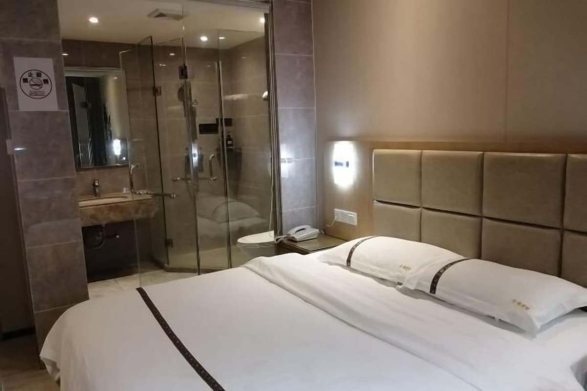 A hotel room with a large bed, a glass-enclosed shower, a bedside table, and a wall-mounted light.