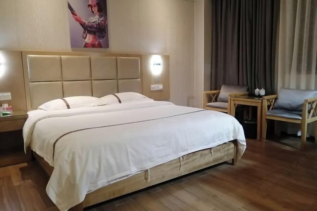 A hotel room with a large bed, a wooden headboard, a nightstand with a lamp, a chair, and a small table with a lamp.