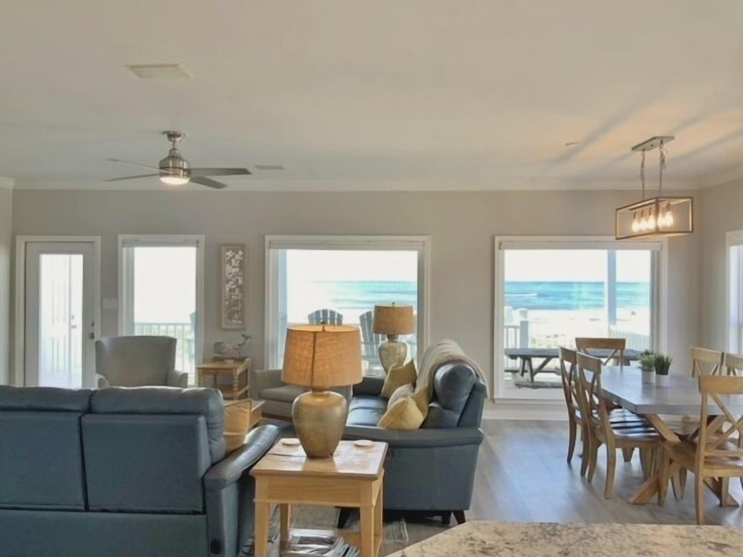 Un salon spacieux avec un grand canapé, un coin repas doté de chaises en bois et une vue sur la plage grâce à de grandes fenêtres.