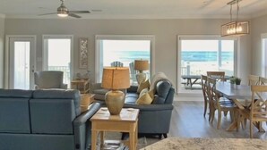 Een ruime woonkamer met een grote bank, een eethoek met houten stoelen en uitzicht op het strand door grote ramen.