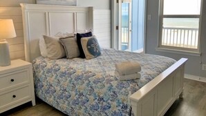 Een slaapkamer met een bed, nachtkastje en uitzicht op het strand door een raam.