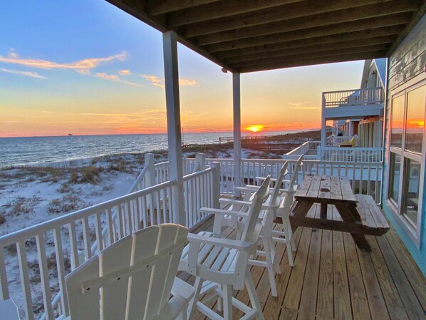A wooden deck with white chairs and a picnic table overlooking a beach at sunset.