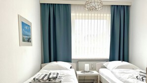 A hotel room with two single beds, a small bedside table with a lamp, a window with blue curtains, and a framed picture on the wall. - Klaus-Groth-Str 2 App 11 Westerland (Westerland)