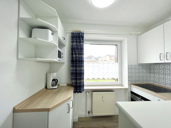 Une cuisine compacte avec des meubles blancs, un plan de travail en bois, un four à micro-ondes encastré et une fenêtre donnant sur des maisons.