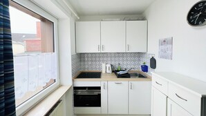 A compact kitchen with white cabinets, a black oven, a sink, and a window with blinds. - Klaus-Groth-Str 2 App 11 Westerland (Westerland)