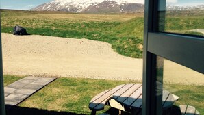 Vista desde el interior de un edificio con mesas y bancos afuera, un campo cubierto de césped y una montaña nevada en la lejanía.