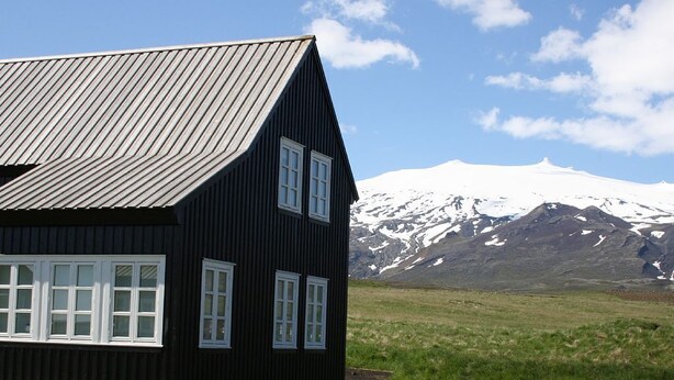 Una casa de madera negra con techo inclinado y ventanas blancas, frente a un paisaje de montaña nevada y cielo despejado.