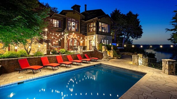 A pool area with red lounge chairs, stone walls, and a view of a lake at dusk. - PRIVATE 10,000 SqFt lakefront log estate  w. Pool  - Golfers Retreat - Close to Big Cedar! (kirbyville)