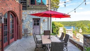A patio with a table and chairs, a red umbrella, and a stone building with a view of a lake. - PRIVATE 10,000 SqFt lakefront log estate  w. Pool  - Golfers Retreat - Close to Big Cedar! (kirbyville)