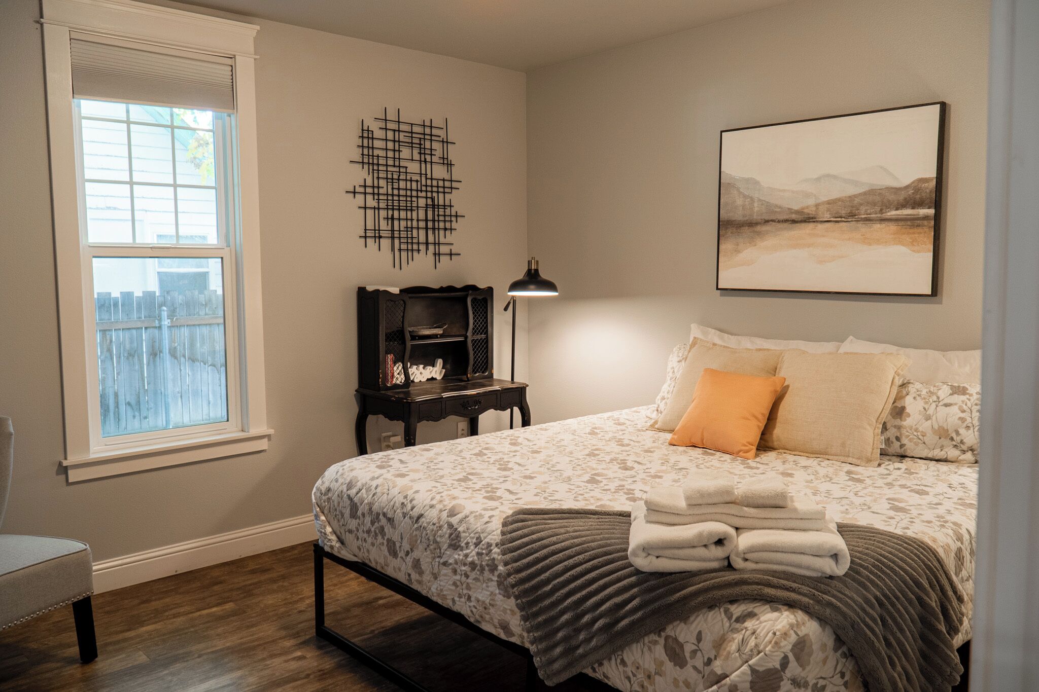 A bedroom with a bed, a nightstand, a lamp, a window, and a wall art piece.