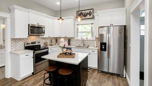 A modern kitchen with white cabinetry, stainless steel appliances, a central island with stools, pendant lighting, and a herringbone tile backsplash. - The Rustic Rose - 4 Bedroom House w/ HOT TUB near Downtown (Walla Walla)