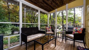 Un porche cubierto con piso de madera, muebles de mimbre negro y amplios ventanales que dan a una zona boscosa.