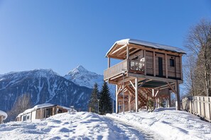 Una cabaña de madera con balcón, con un paisaje nevado de montañas al fondo.