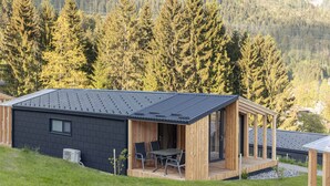 Una baita moderna con tetto scuro, terrazza in legno e zona pranzo con sedie in metallo.