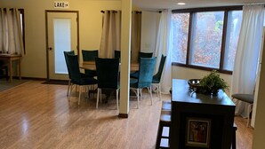 Un'ampia stanza con pavimento in legno, una ventola a soffitto, un'area pranzo con sedie, un tavolino con una pianta e una porta con la scritta 'LAKE'.