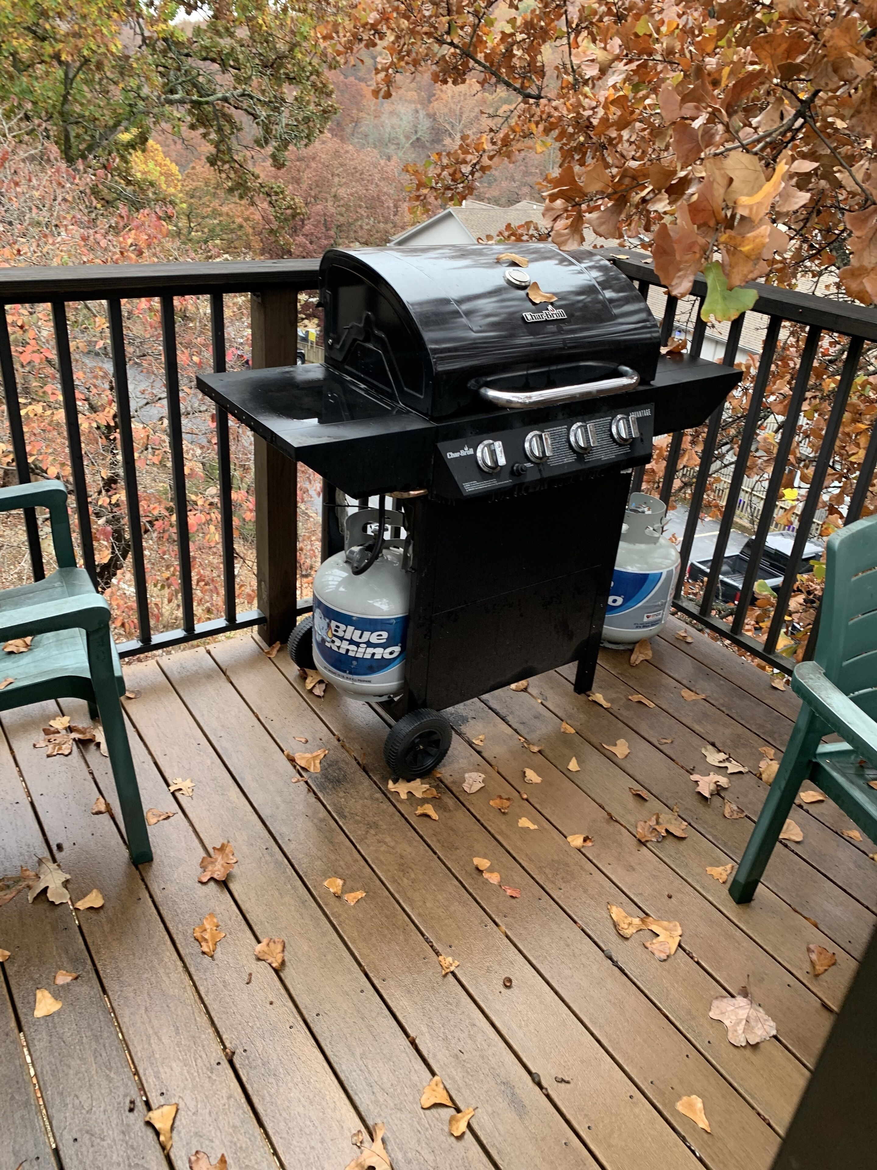 A portable grill on a wooden deck with autumn leaves, two green chairs, and a gas tank labeled "Blue Rhino".