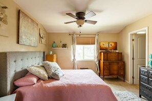 A bedroom with a bed, a ceiling fan, a window with curtains, a dresser, and artwork on the walls.