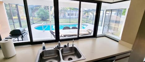 A kitchen with a large window overlooking a backyard with a pool and outdoor furniture.