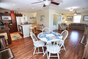 A spacious living area with a dining table surrounded by white chairs, a kitchen with wooden cabinets and a microwave, and a ceiling fan.