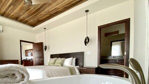 Een hotelkamer met een groot bad, een bed met witte lakens, een houten plafond en een keukenruimte zichtbaar door een geopende deur.