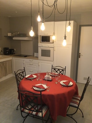Un coin repas avec une table ronde dressée pour quatre personnes, entourée de chaises dotées de coussins à rayures. Au-dessus de la table, on peut voir des ampoules suspendues. La pièce est équipée de meubles blancs et un four à micro-ondes est posé sur le plan de travail.