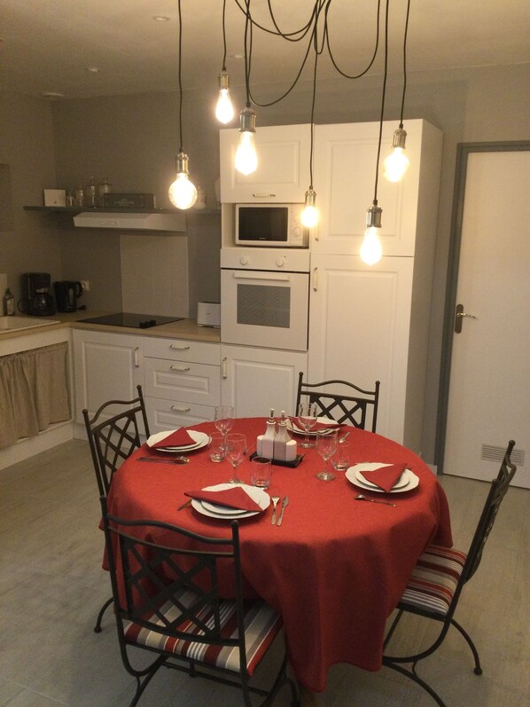 Un coin repas avec une table ronde dressée pour quatre personnes, entourée de chaises dotées de coussins à rayures. Au-dessus de la table, on peut voir des ampoules suspendues. La pièce est équipée de meubles blancs et un four à micro-ondes est posé sur le plan de travail.