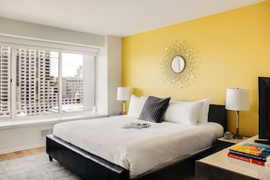 Quarto de hotel com uma cama grande, parede amarela de destaque, espelho redondo e vista para edifícios altos.