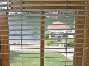 Ein Fenster mit horizontalen Rollläden, Blick auf eine grüne Rasenfläche und ein Gebäude mit rotem Dach im Hintergrund.