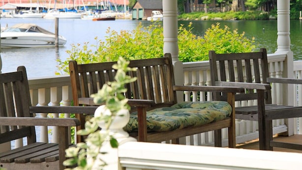 A wooden bench with a cushion, a white balustrade, and a view of a marina with boats and a dock. - Waterfront Smith Cove Lake House includes dock (Gilford)