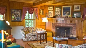 A cozy living room with wooden walls, a fireplace, floral-patterned sofas, and a green armchair. - Waterfront Smith Cove Lake House includes dock (Gilford)