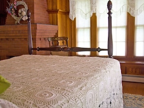 Ein Bett mit hölzernem Kopfteil, einer Spitzenüberdecke und einem Korbstuhl in einem Zimmer mit Holzwänden und Fenstern mit Spitzenvorhängen.