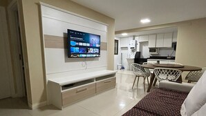 A modern living room with a flat-screen TV mounted on the wall, a dining area with a table and chairs, and a kitchen in the background. - Arraial Praia dos Anjos Residence Club (Arraial do Cabo)