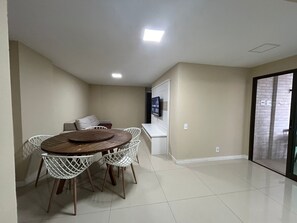 A dining area with a round table and white chairs, a sofa, and a TV mounted on the wall. - Arraial Praia dos Anjos Residence Club (Arraial do Cabo)